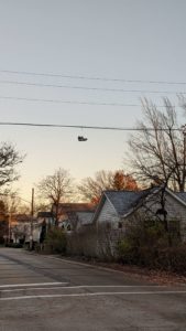 A street with a row of modern houses on the right side. A powerline goes across the street with a pair of grey nike shoes hanging from it. There is a gentle sunset in the cloudless sky.