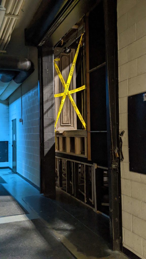 A white brick hallway that is bathed in a blue light. At the center, there is an open window into another room. Laid over the opening are three pieces of yellow tape labelled "Caution" in an asterisk pattern.