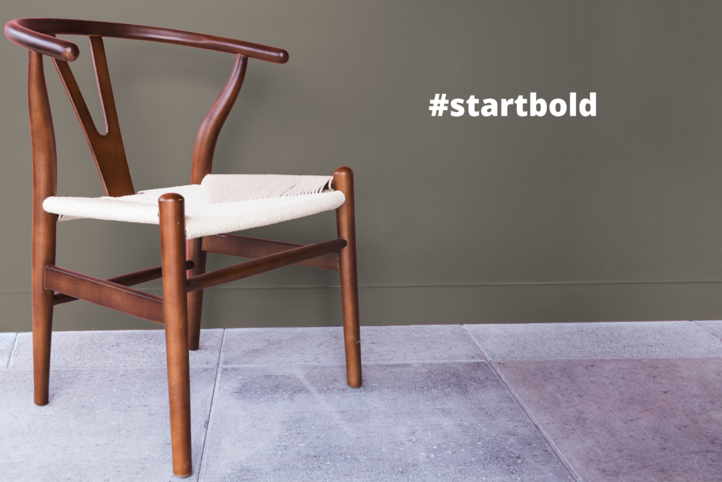A wooden chair on a stone ground against a green background. To the right of the chair is the text #startbold.